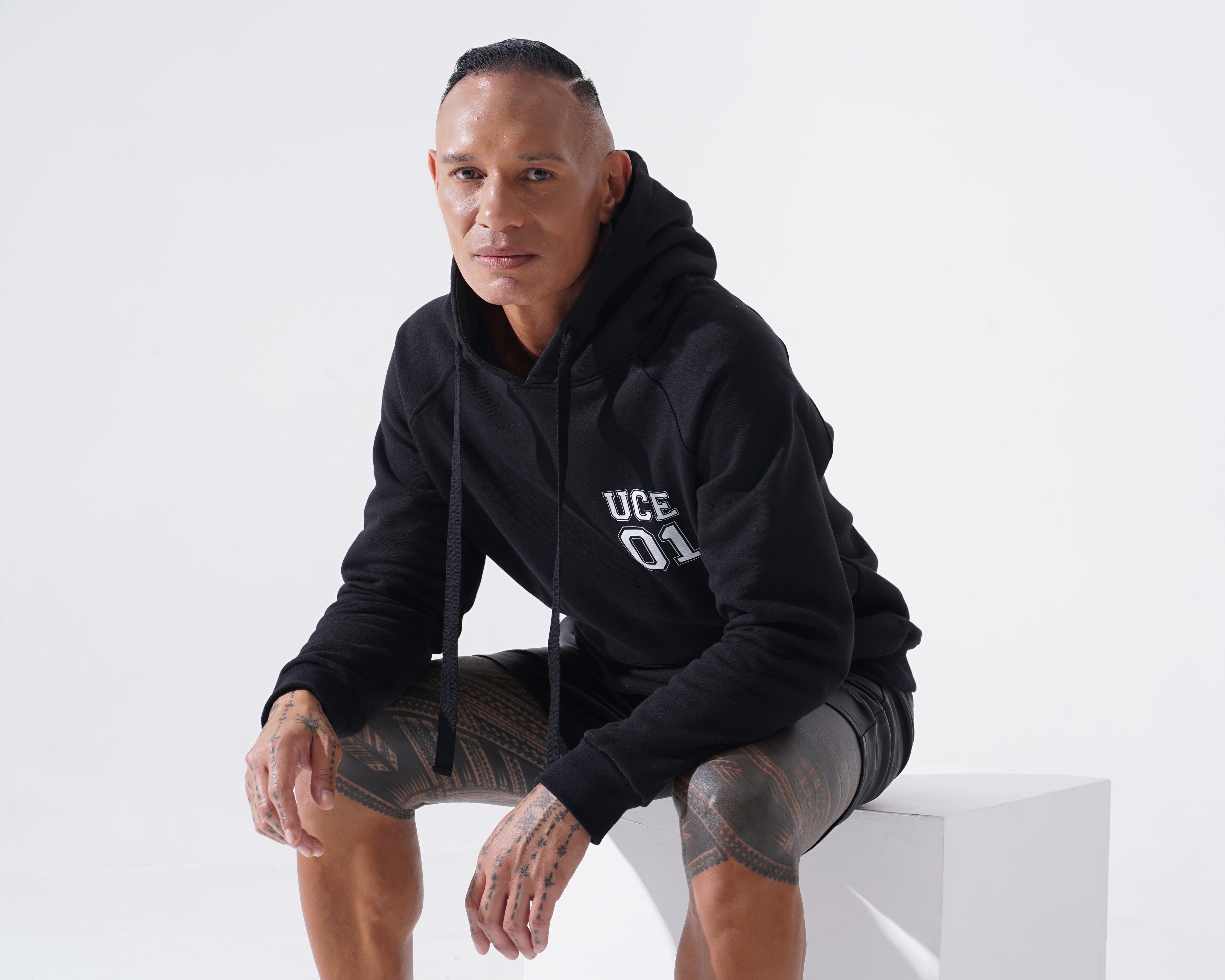 Man wearing a black hoodie with text, sitting on a white chair against a white background